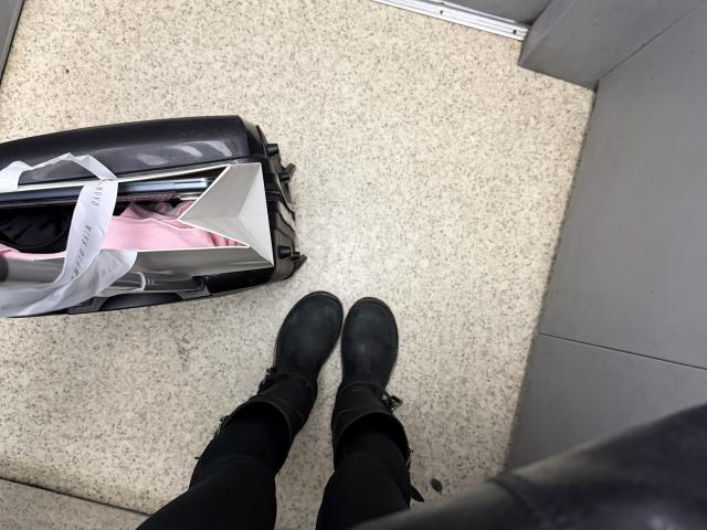 Les bottes noires de SensualMila se trouvent sur un sol moucheté, avec une valise noire ouverte, montrant des vêtements roses et des étiquettes blanches, dans une pièce aux murs gris..