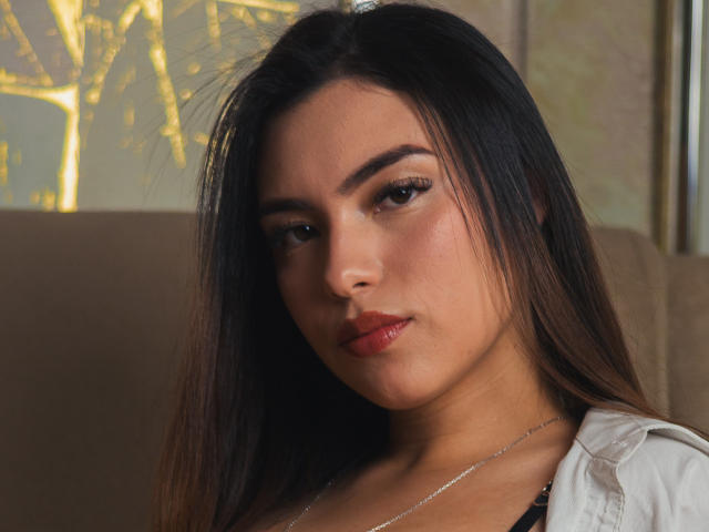 CelesteVelasco with dark straight hair, brown eyes, and red lipstick, wearing a white shirt, showing a slight pout, and a silver necklace, looking directly at the camera.