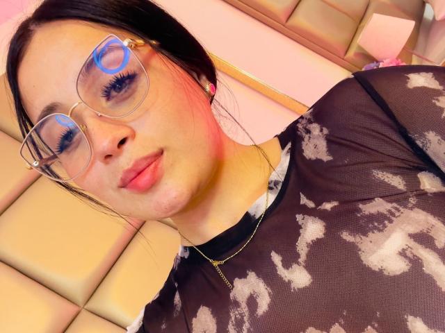 AnnyBentleii, usando óculos, com lábios rosados e uma blusa preta transparente que mostra os seios, usa um colar dourado e tem a pele rosada, olhando para a câmera com uma expressão levemente emburrada..