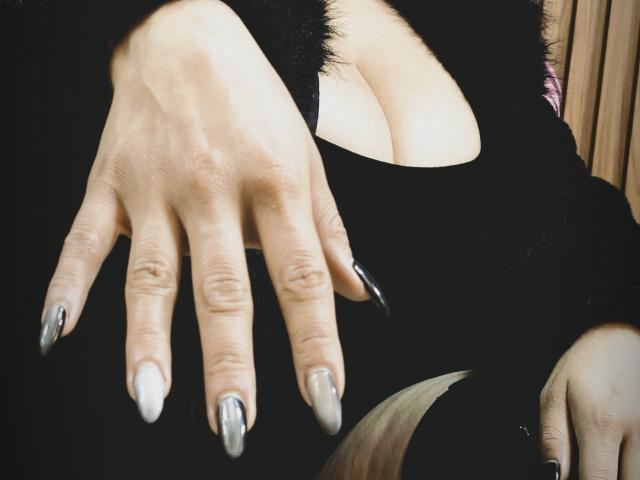 De hand van Barbara Leythonn, met lange, zilveren, puntige nagels, rust op haar borsten, bedekt met een zwart, pluizig topje met uitsparingen, dat een beetje decolleté laat zien. Haar huid is licht..