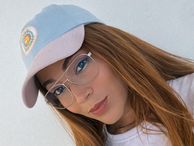EmmaLym, con el pelo largo y rojo, lleva puesto un gorro azul claro con un emblema colorido y gafas transparentes. Tiene un piercing en la nariz que le queda lindo y está mirando ligeramente hacia abajo..