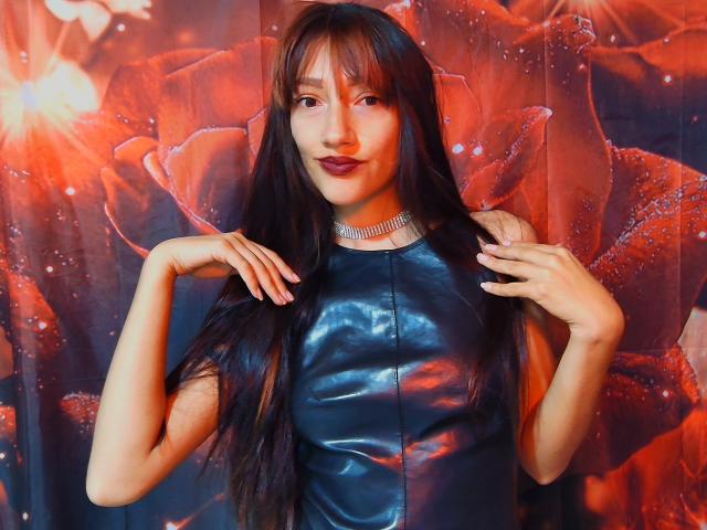 KiriamGomez met lang zwart haar, gekleed in een glanzende blauwe leren jurk, die haar haar vasthoudt, met een glimlach en donkere lipstick, en een sprankelende choker. Rode bloemen behang op de achtergrond..