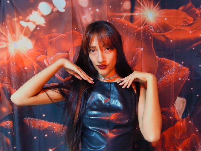 KiriamGomez with long brown hair, wearing a shiny black leather top, has hands near face, dark lipstick, and a necklace, set against a vibrant red flower background.