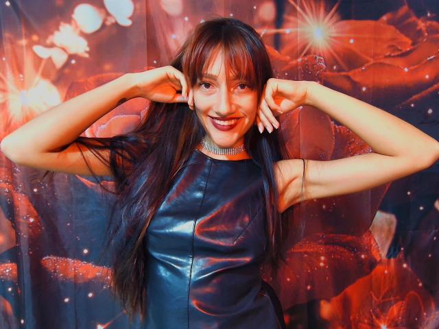 KiriamGomez with long brown hair, smiling, wearing a black shiny leather dress, hands in hair, sparkling choker, red and orange bokeh background, looking cute, sexy, and confident.