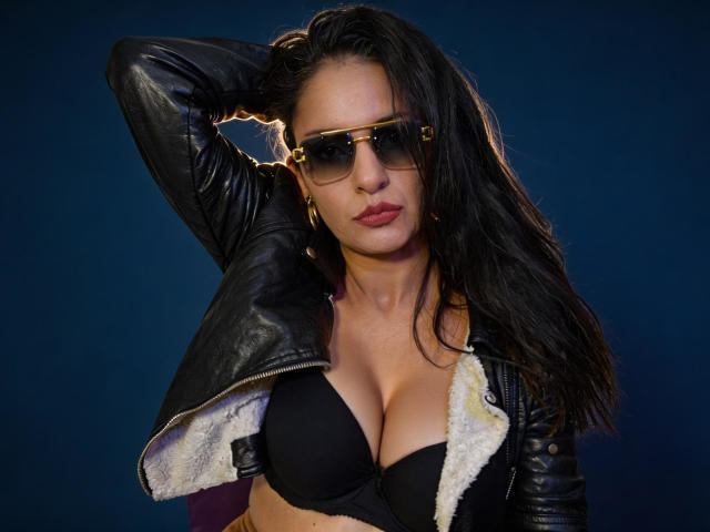 AngelaBranson with dark hair, black leather jacket, and sunglasses, showing her black bra-covered boobs, hand in hair, looking serious, against a dark blue background.