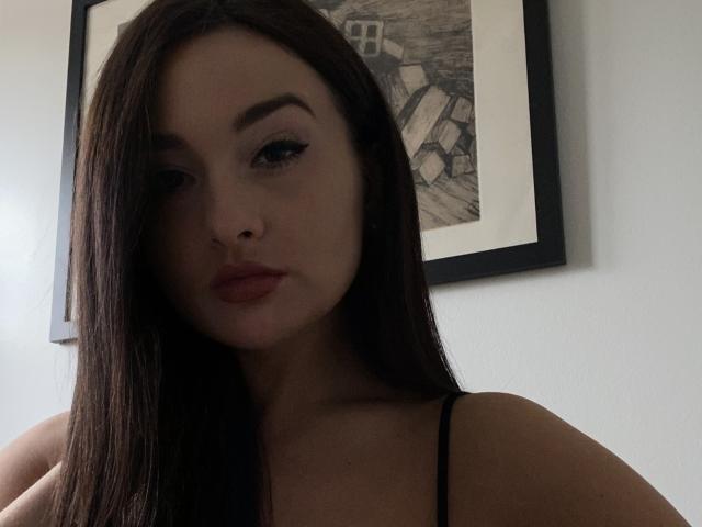Dionisia ha lunghi capelli castano scuro, sopracciglia folte e rossetto rosso. Indossa una canotta nera a spalline, con il seno leggermente visibile. La sua espressione è neutra e seria..