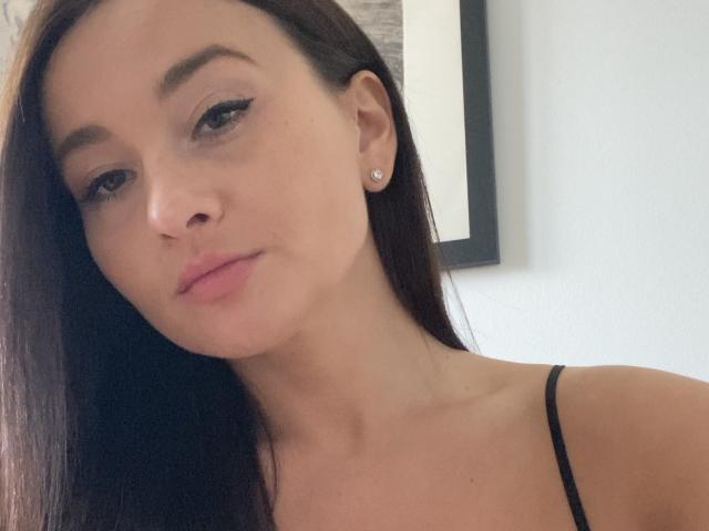 Dionisia has long brown hair, subtle makeup, and a slight smile. She's wearing a black spaghetti strap top with small diamond earrings, and her tits are barely visible.