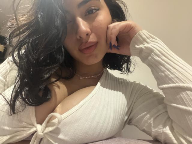 EmmaBeaumont with wavy black hair, pouty lips, and blue nails, wearing a white ribbed top with a deep cleavage showing her big tits, resting her chin on her hand.