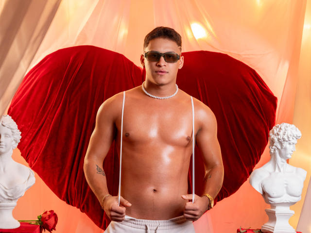 Shirtless JamesOenn with red velvet wings, wearing sunglasses, necklace, and white pants, holding suspenders, standing between white statues, tan, muscular, tattoo on left arm.