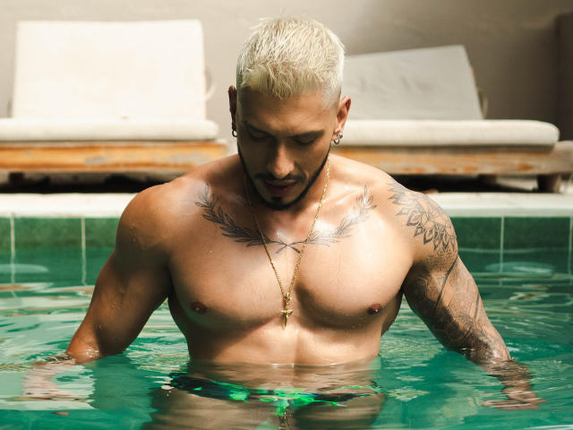 SethVegas with bleached hair, tattoos on chest and arm, and a beard, standing in a pool, looking down, wearing a gold necklace, with green water covering his ass.