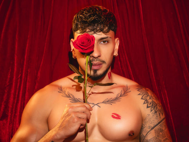 SethVegas with a rose in front of his face, holding it with one hand. He's shirtless, has tattoos, a lip kiss mark, and red rose tattoo on his chest.