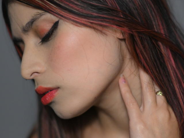 TifanySuarez has red lips, closed eyes with black eyeliner, red eyeshadow, and dark red-black hair. Her hand touches her neck, showing a green ring.