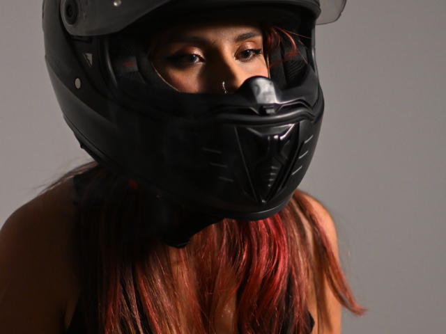 TifanySuarez with dark brown skin, wearing a black motorcycle helmet, and red-tinted long hair, covering her nose and mouth, looking through the visor.
