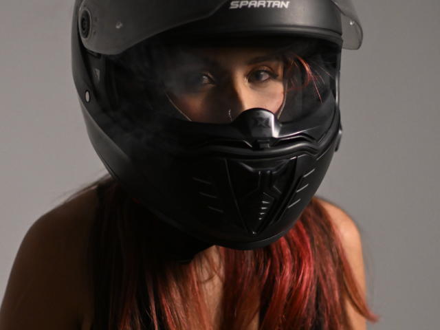 TifanySuarez with black Sparatan helmet, showing only eyes and nose, has red hair, and a slight smirk. Helmet obscures mouth and chin, giving a mysterious look.