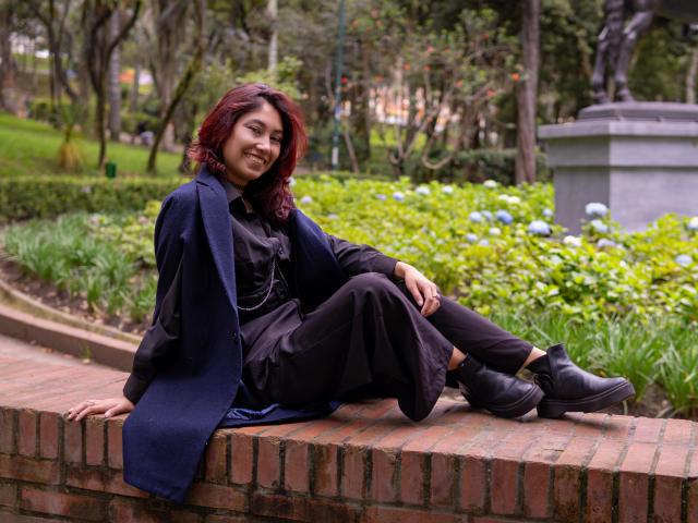AliCipher with red hair, smiling, sitting on brick ledge, wearing dark dress, black pants, black boots, navy blue scarf, hands on brick, legs crossed, relaxed, green garden.