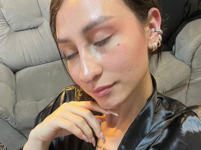 MaddyCarina with shiny skin, brown hair, and a black leather jacket, touching her chin with long, manicured nails. She's wearing ornate earrings and has a gentle, closed-eye smile.