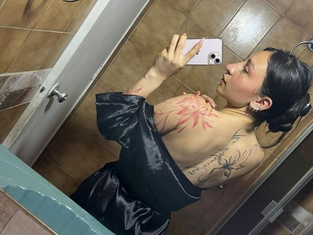 MaddyCarina mit Tattoos auf dem Rücken und Arm, schwarzes Hemd ausgezogen, macht ein Selfie vor einem Spiegel in einem Badezimmer, hält ein weißes Telefon, braune Fliesenwände, kurze schwarze Haare..