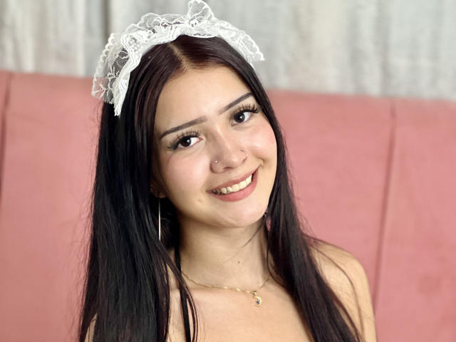 KarinaCurvy has long black hair, wearing a white lace headband, smiling with her teeth showing, has a small nose ring, and a gold necklace, with pink background.