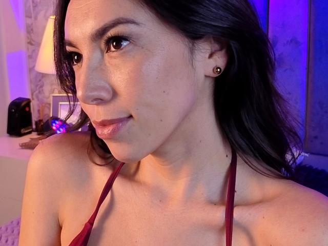 MiaReed with shiny skin, dark hair, small earrings, and a red halter top, smiling slightly, brown eyes, subtle makeup, and small tits, in a softly lit room.