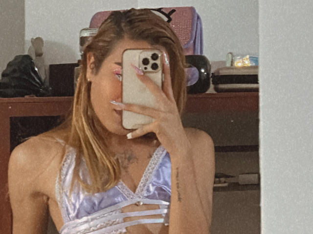 EmmaLym, con cabello rubio, vistiendo un sujetador morado brillante, tomando una selfie frente al espejo con su teléfono, mostrando un tatuaje en su pecho y uñas largas..
