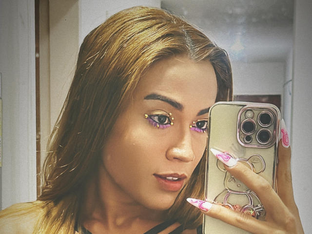 EmmaLym, con piel morena, cabello castaño y maquillaje de ojos morado, sosteniendo un teléfono con una funda rosa con forma de oso, mostrando uñas largas y rosas, en un selfie frente al espejo..