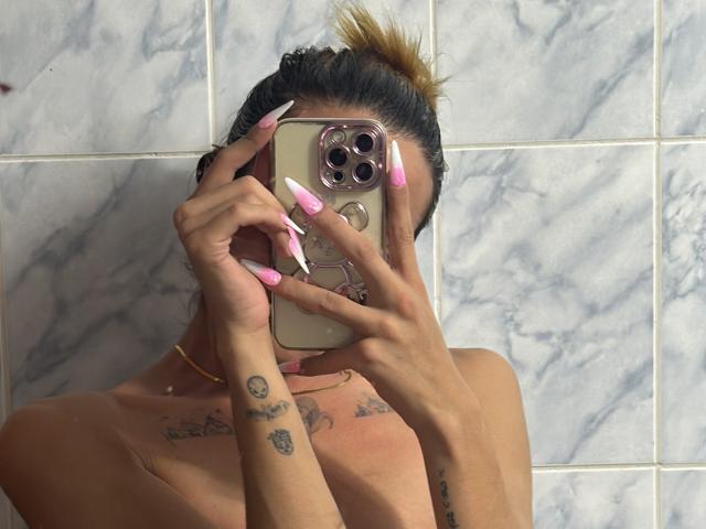 EmmaLym with tan skin, pink and white long nails, holding a phone covering her face, topless, tattoos on hands and chest, brown hair in a bun, marble tiled wall.
