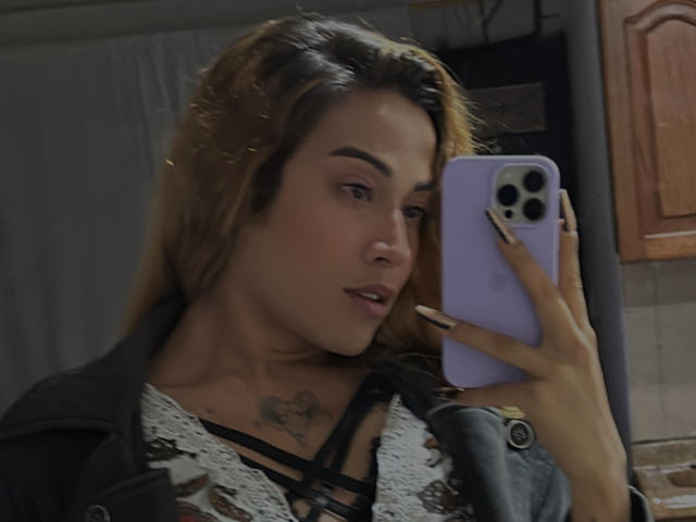 EmmaLym, met lichtbruin haar, die een paarse telefoon vasthoudt, een zwarte top met veters draagt, een tatoeage op haar borst laat zien, en naar de telefoon kijkt, in een slecht verlichte kamer..