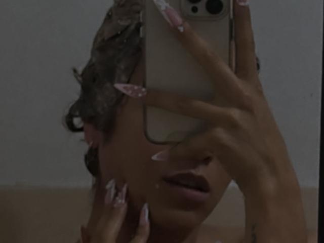 EmmaLym che si fa un selfie, con il telefono che le copre il viso, mostrando capelli ricci scuri, unghie rosa e la bocca leggermente aperta, in una stanza poco illuminata..