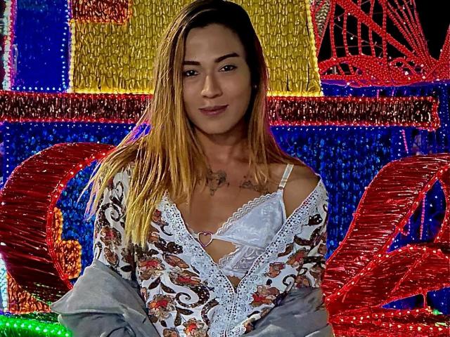 EmmaLym, met lang bruin haar, draagt een witte bustier met veters die haar kleine borsten laat zien, en een top met een bloemenprint, en ze glimlacht naar de camera. Ze heeft tatoeages op haar borst..