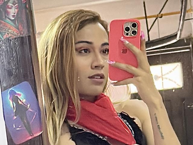 EmmaLym, avec ses cheveux blonds mouillés, ses ongles roses, son foulard rouge, son haut noir, tenant un téléphone rose, se prend en photo devant un miroir, avec un petit piercing au nez, un tatouage sur l'avant-bras droit, du rouge à lèvres rose et un sourire confiant..