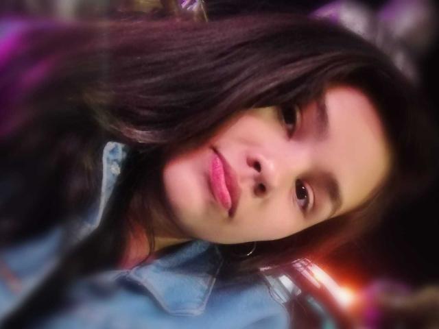 RubyCrow has dark, wavy hair, pink lips, and a slight smile. She wears a blue denim shirt, with blurry purple and orange lights in the background.