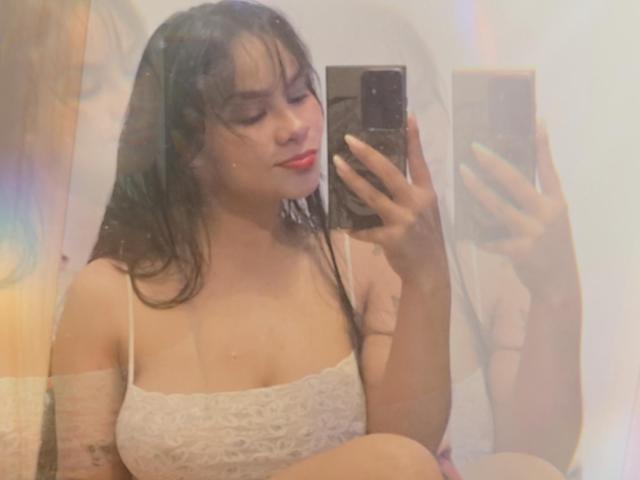Yurimy with wet black hair, red lipstick, white lace top, holding phone, taking selfie, showing her tits, sitting, smiling, holding phone in mirror.