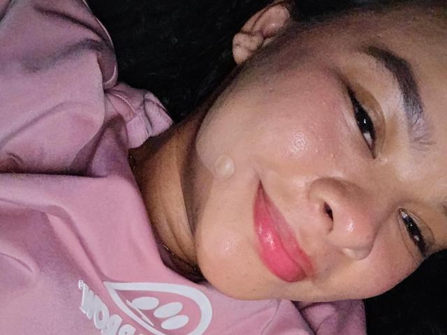 O rosto de Yurimy, suado com uma pequena gota na bochecha, vestindo uma camisa rosa, sorrindo suavemente com lábios rosados, deitada, close-up, fundo escuro..