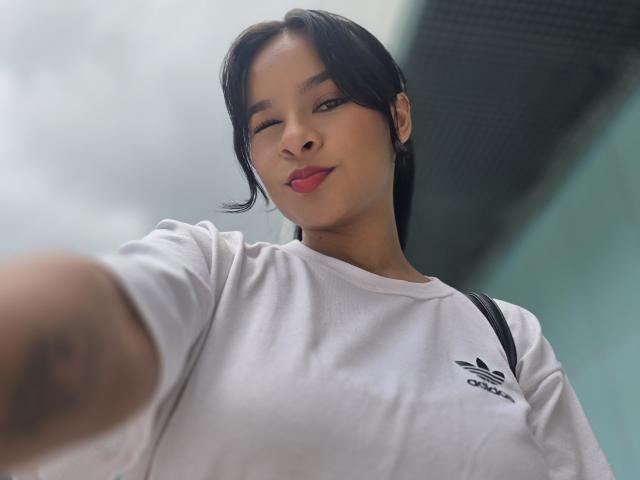 Yurimy with short black hair, winking, wearing a white Adidas shirt, has a tattoo on her right arm, and is taking a selfie, her red lips slightly parted.
