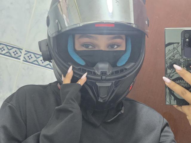 Yurimy usando um capacete preto com interior azul, uma máscara preta e uma camisa preta, segurando um telefone com unhas bem cuidadas, tirando uma selfie, mostrando apenas seus olhos..