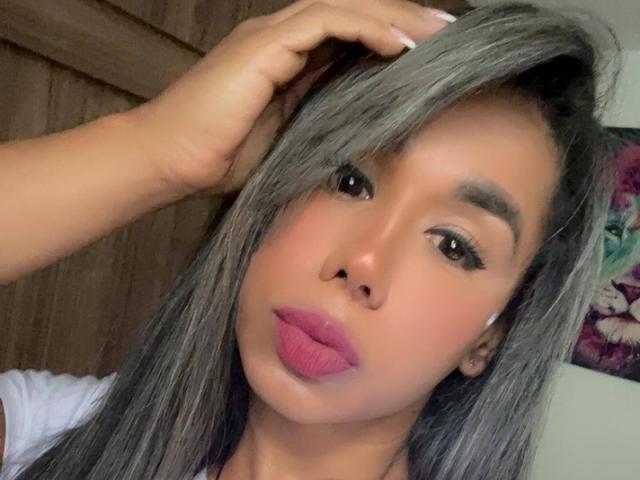 Iria with gray hair, pink lipstick, and brown eyes, hand in hair, looking at camera, showing off her pouty ass-lickable lips.