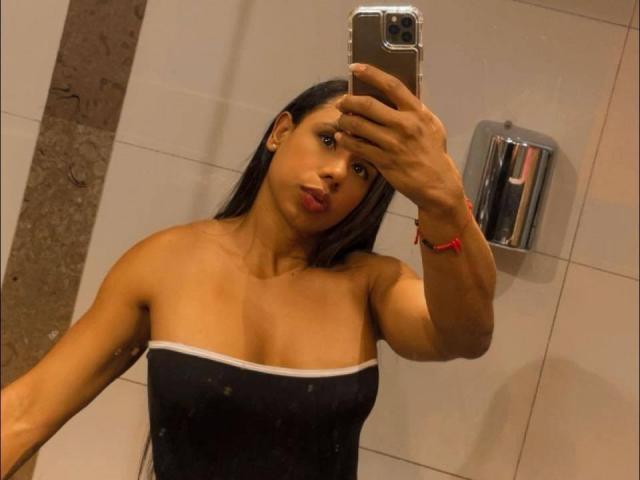 Iria with medium brown skin, long black hair, and pouty lips, wearing a black strapless top, taking a mirror selfie with her right hand, red bracelet on wrist.