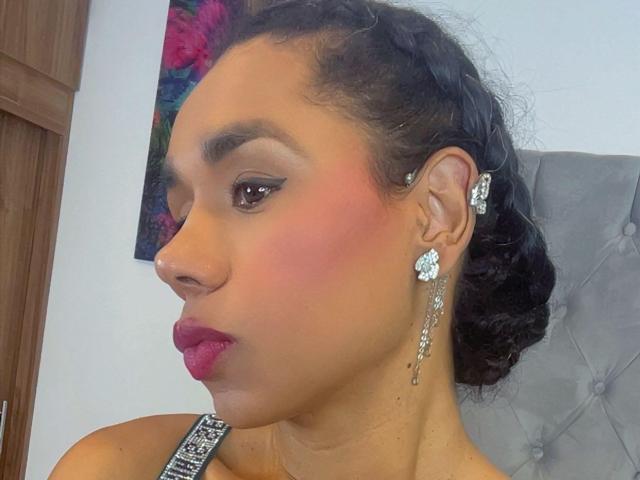 Iria has brown skin, curly black hair in a bun, pink blush, red lipstick, and wears diamond earrings with matching studs. She's in profile, looking down.