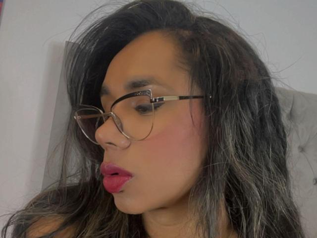 Iria with dark, messy hair, wearing glasses, has pink blush and red lipstick, pouting her glossy lips. Her tan skin is smooth, and she's looking slightly to the left.