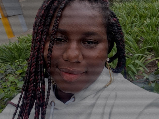 Dilay with dark skin and red-black braided hair, wearing a white shirt, smiling subtly, has small gold hoop earrings, and a slightly shiny face, looks relaxed.