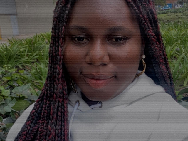 Dilay's dark brown face with red and black braids, wearing a white hoodie, has a subtle smile with a small black mark under her left eye. Her gold hoop earring.
