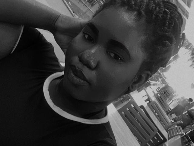 Black and white selfie of Dilay with dark skin, curly hair, and a pensive expression, resting their head on their hand. Dilay's nose has a small freckle.