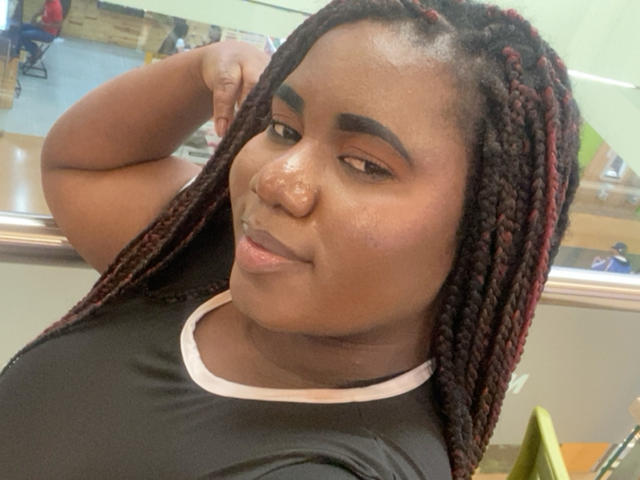 Dilay with dark brown skin, long red and black braided hair, small nose piercing, black shirt with white trim, smiling slightly, arm raised behind head, looking at camera.
