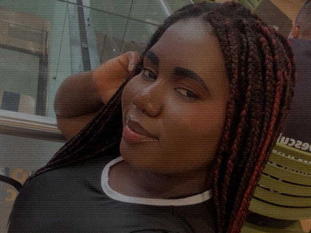 Dilay's dark brown face with red and black braided hair, wearing a black shirt with white trim, smiling slightly with a hand near her head.