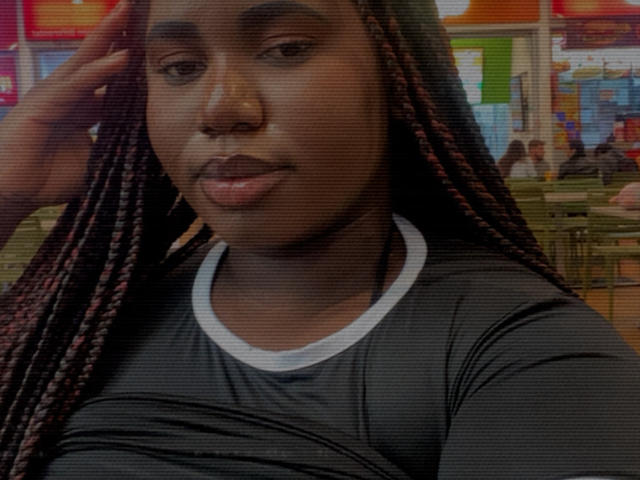 Dilay has dark skin, long red-brown braids, and wears a black shirt with a white neckline. She's looking at the camera with a neutral expression.