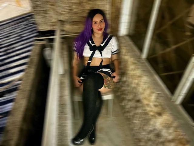 Amy Ross, con cabello morado, lápiz labial rojo, una blusa de marinero en blanco y negro, una falda negra, medias negras hasta el muslo y tacones negros, sentada en una silla, mostrando tatuajes en su muslo izquierdo..