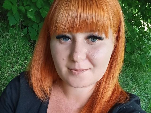 Kirobra69 has bright orange hair, blue eyes with thick black eyeliner, and a light skin with freckles. She's wearing a black top, smiling slightly, and has a small chin scar.