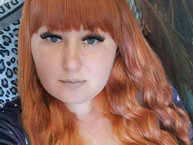 Kirobra69 tem cabelo ruivo, olhos azuis e usa delineador preto. Seu nariz é pequeno e ela tem uma expressão neutra com batom marrom claro. Ela está olhando para a câmera..