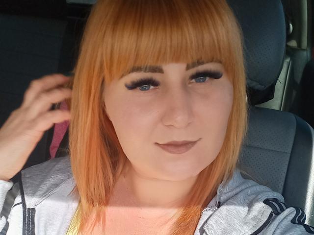 Kirobra69 hat orangefarbenes Haar, blaue Augen und trägt dunkle Make-up mit dicken Wimpern. Sie sitzt in einem Auto, trägt eine graue Jacke und hat eine Hand in ihrem Haar..