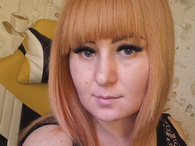 Kirobra69 has light orange hair, blue eyes, and thick black eyelashes. She's looking at the camera with a slight smile, wearing a black lace top.